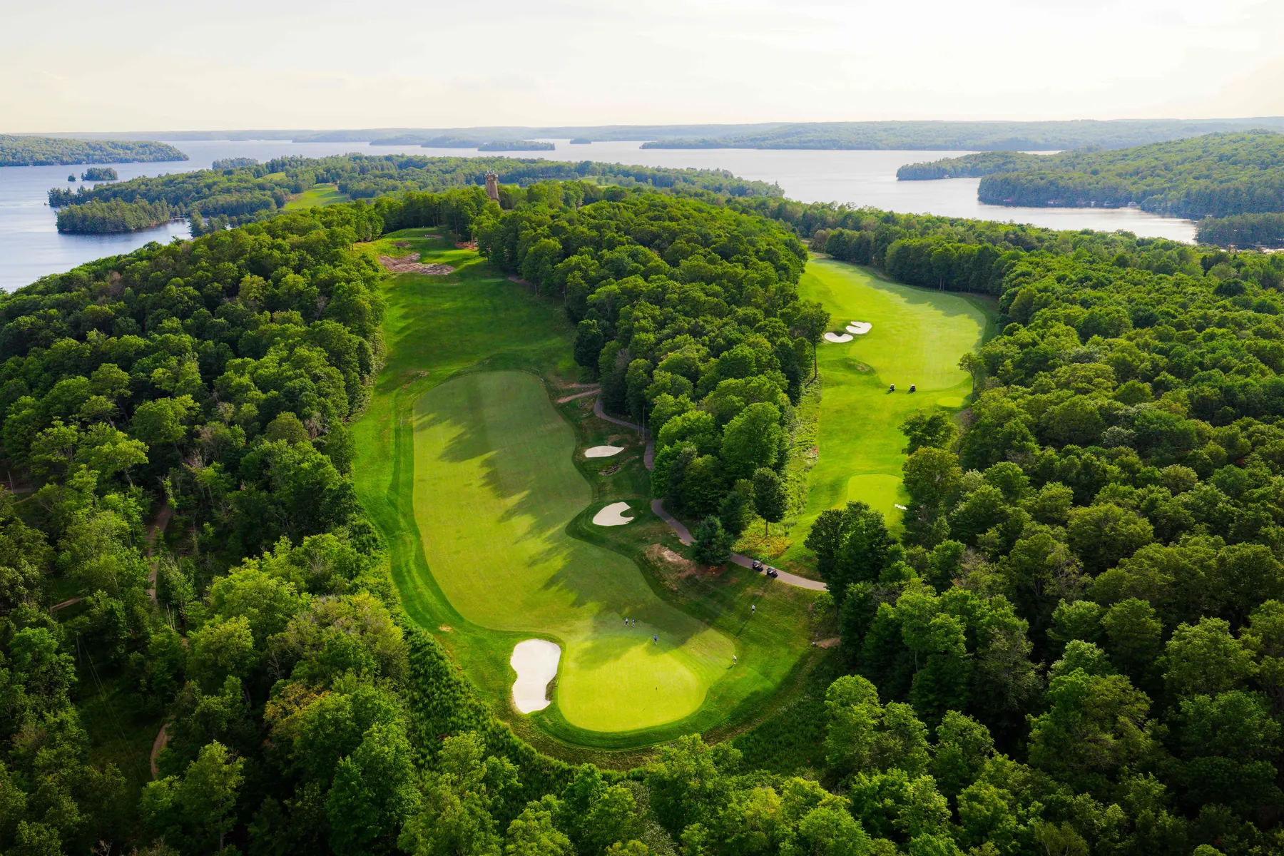 Aerial of Bigwin Island's fairways through the forest, with the stone tower and Lake of Bays beyond